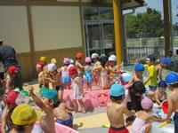 赤、白、黄色、青、水色の色とりどりの水泳帽をかぶった園児が楽しそうに園庭でプール遊びをしている写真
