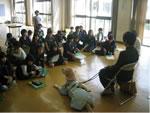 盲導犬が座りその横に椅子にすわった人の話を床にすわり聞いている生徒達の写真