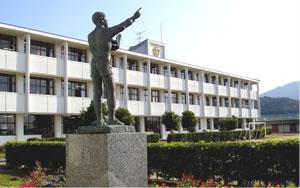 秦荘東小学校の校舎を背景に銅像が右手を挙げて指を指している写真
