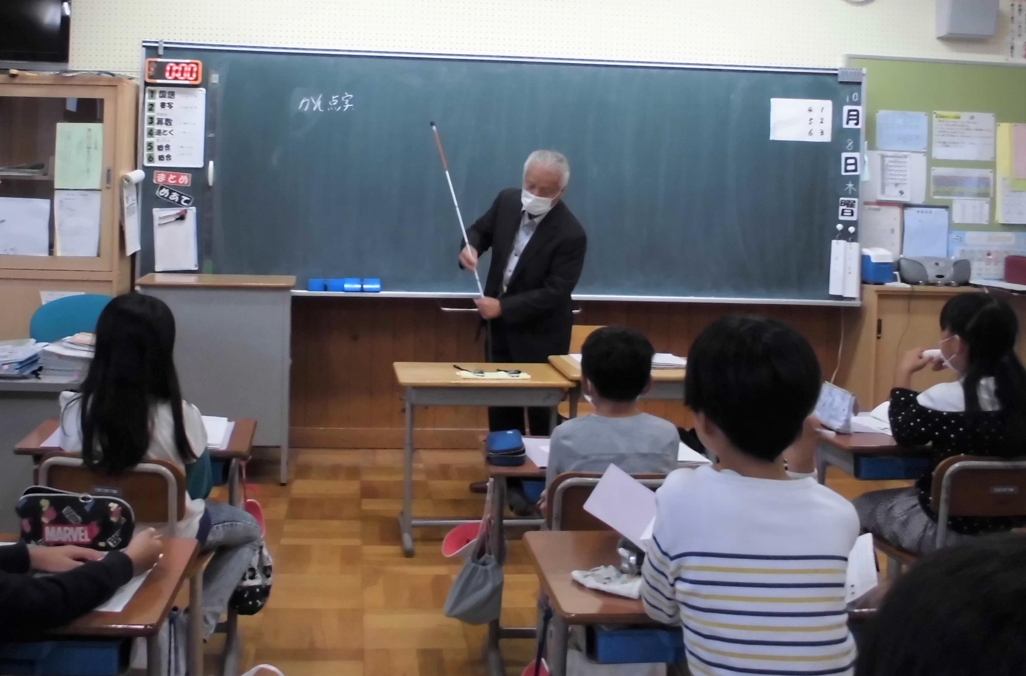 浅野先生が白杖を子どもたちに見せながら、使い方を説明しています。
