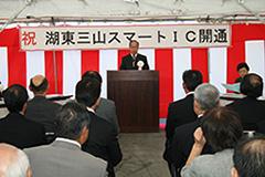 紅白幕の前で挨拶をしている村西町長と記念行事に参加している人達の後ろから撮った写真